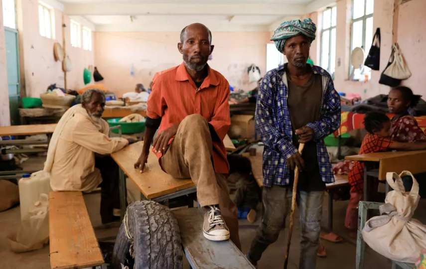 A photo of two men looking at camera inside sit inside their shelter at the Abi Adi camp for the Internally Displaced Persons in Tigray Region, Ethiopia, with a few people behind them.