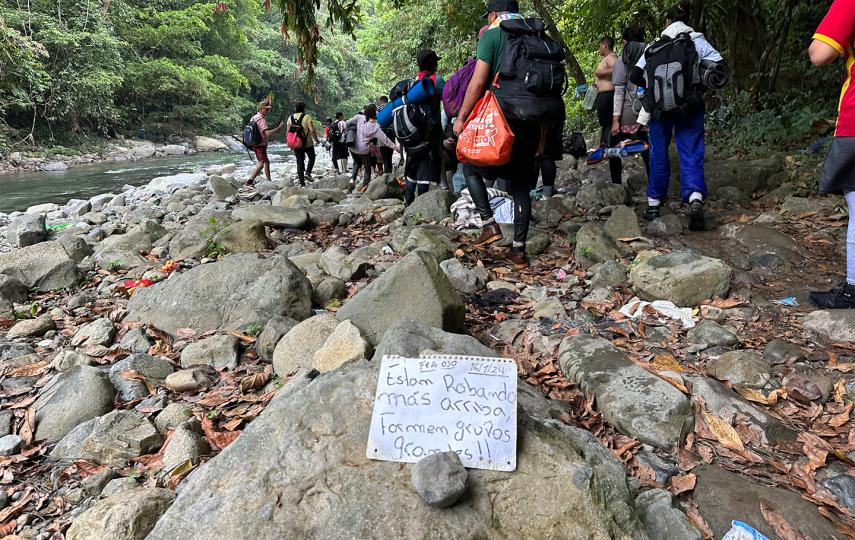 In the foreground we see a paper sign left by migrants warning others of gangs of thieves. The sign reads: “Están robando más arriba, formen grupos grandes!!”/“They're robbing further up, form big groups!!”. In the background we see a group of migrants walking.