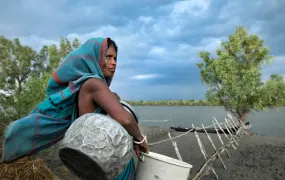 This is a wideshot portrait of Dipu Bala. She turns her back towards the camera and looks to the right of the frame. She holds a metal jug in between her right arm and waist and a white bucket with her right hand. In the background we see a tree and a body of water.