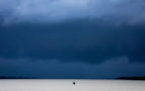 This is a wide shot showing a boatman as they hurry across a river in Khulna District as ominous rain clouds loom in the sky. 