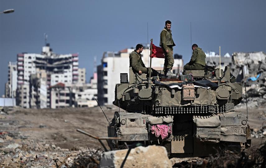 This is a wide shot showing a military tank with three soldiers on it. One stands while the other two sit. In the back you can see a skyline in the Gaza strip along with rubble.