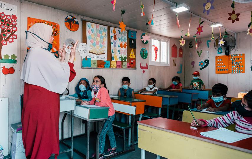 Pictured is a teacher standing in front of a class of children and gesturing as she is speaking to them. The children are sitting and looking at her. Everyone is wearing a medical facemask. The classroom is colorfully decorated.